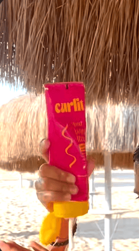 Bright pink Curlit hair curling product in a tube, held under a beach umbrella on a sunny day, emphasizing beach-ready, easy-to-use curling solutions for perfect waves.